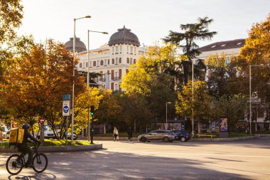 Madrid, İspanya 22 Kasım 2023 güneşli sonbahar gününde bir şehir caddesi. Bisikletçi, Glovo teslimat servisi kuryesi yol boyunca gidiyor. Şehir manzarası. Şehir caddesindeki arabalar. Güzel klasik mimari