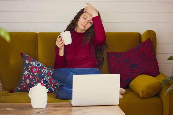 A young beautiful woman with long curly hair is relaxing at home sitting on a brown sofa with a white cup of tea in her hands watching an interesting comedy movie. A quiet, relaxed girl in a cozy room