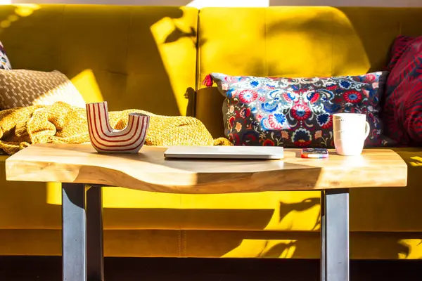 Modern interior in a Scandinavian style. Yellow sofa and a little wooden table with vase and cup in cozy home full of light. A house from inside with designer elements. Multicolored decorative pillows