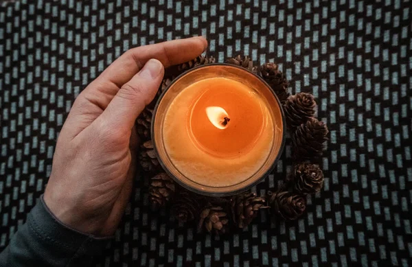 Yellow round candle in a woman\'s hand on a dark ornamental background flatly top view. Home decor details. Woman holding a scented aromatic burning candle among pine cones. Touch a fire. Warm light.