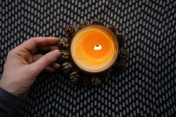 Yellow round candle in a woman\'s hand on a dark ornamental background flatly top view. Home decor details. Woman holding a scented aromatic burning candle among pine cones. Touch a fire. Warm light.
