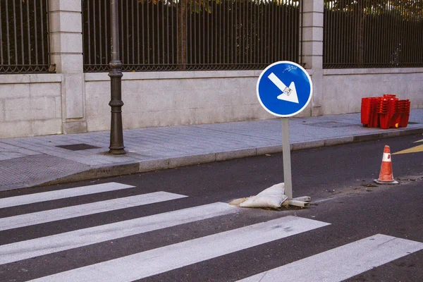 Painted pedestrian zebra crossing in a city. A road round blue sign with arrow. Renovation work in an urban environment. Cityscape landscape. Red traffic cone barrier. Construction elements in town.