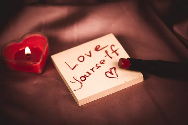 A note to self with a call to love yourself drawing with red lipstick on a silk dark pink cloth flatly. A burning heart-shaped red candle, gift box. Happy Valentines Day 2024. Love yourself first.
