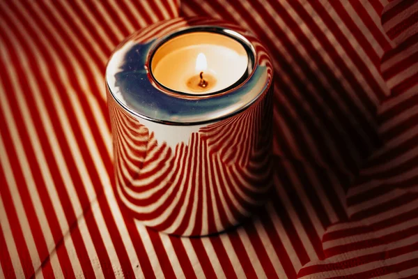 A burning candle in a metal candle holder on a striped red and white background. A candle glows in the darkness. Wax candle in a stylish modern holder on a table in a dark room. Interior details.