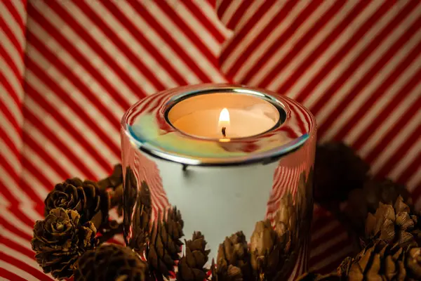 A burning candle in metal candle holder on striped red white background. A candle glows in a darkness. Wax candle in stylish modern holder on table in dark room. Interior details. Christmas tree cones