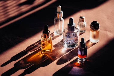 Farklı kozmetik ürünlerinden oluşan estetik cam şişeler pembe kozmetik masasının üzerine uzun gölgeler bırakıyor. Serum şişesi ve pipet, parfüm, tırnak yağı..