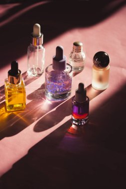 Farklı kozmetik ürünlerinden oluşan estetik cam şişeler pembe kozmetik masasının üzerine uzun gölgeler bırakıyor. Serum şişesi ve pipet, parfüm, tırnak yağı..