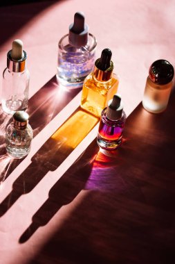 Farklı kozmetik ürünlerinden oluşan estetik cam şişeler pembe kozmetik masasının üzerine uzun gölgeler bırakıyor. Serum şişesi ve pipet, parfüm, tırnak yağı..