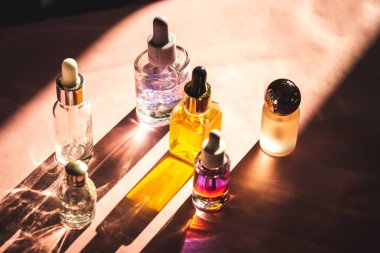 Farklı kozmetik ürünlerinden oluşan estetik cam şişeler pembe kozmetik masasının üzerine uzun gölgeler bırakıyor. Serum şişesi ve pipet, parfüm, tırnak yağı..