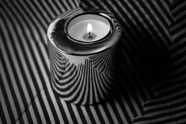 A burning candle in a metal candle holder on a striped surface. Black and white photo. A candle glows in a darkness. Wax candle holder in a darkness.