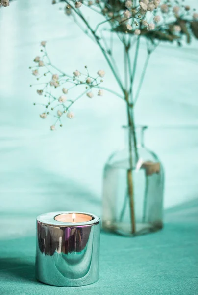 A burning candle in metal candle holder on blue table. Stylish decor for home interior. Still life with a glass vase and small white blooming flowers.