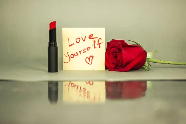 Red lipstick and a note with hand written text \'Love yourself\' and a heart shaped draw on a glass mirror cosmetic table. Motivation to love yourself.