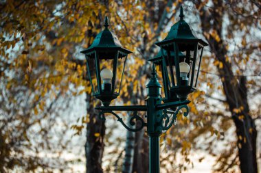 Dekoratif retro dökme demir fener ve sonbahar parkında üç lamba. Sonbaharda dışarıda antik bir lamba direği var. Güz mevsiminde, sarı yapraklı ağaçlara karşı güzel bir sokak lambası. Karanlık doğa fotoğrafı. 