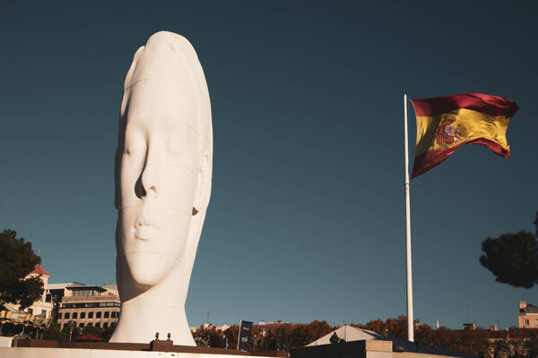 Madrid, Spain. November 22 2023 High huge white sculpture of young woman with eyes closed against blue sky. Masterpiece by artist Jaume Plensa on Colon square. Spanish flag on a wind. Dreamy girl face