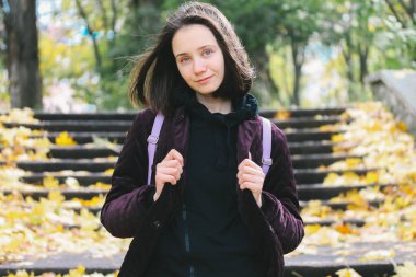Genç, güzel, esmer, Ukraynalı mavi gözlü kadın sonbahar parkında yürüyüş yapıyor, sonbahar mevsimi doğasında. Sırt çantalı mutlu öğrenci kız. Gezgin.