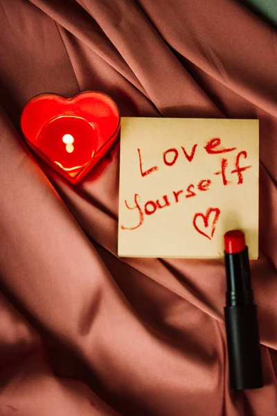 A note with text love yourself drawing with red lipstick on a silk terracotta silk. A burning heart-shaped red candle, home decor. Valentine\'s Day.