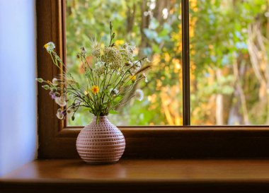 Yazın güneşli bir günde, pencerenin kenarındaki pembe bir vazoda farklı kır çiçeklerinden oluşan bir buket. Evin iç kısımlarında çiçekler var. Rahat bir ev ortamı. Mesaj için yer. Doğal arkaplan.