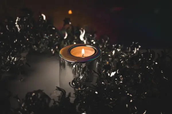 Burning candle in metal candle holder amongst silver tinsel for Christmas tree on dark background. Fire, candle flame. Festive party decor.