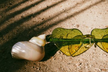 Yeşil kalp şeklinde güneş gözlüğü, kumlu deniz kabuğu. Yazın tropik tatil köyünde okyanus kenarında tatil. Palmiye ağaçları soyut dikey gölgeler.