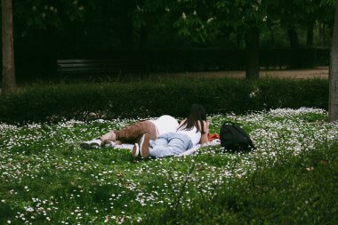 Madrid, İspanya. 1 Mayıs 2024 Bir çift adam ve bir kız baharda, bahçede çiçek açan çiçeklerle yeşil çimlerde yatıyorlar. Dışarıda dinleniyorum.