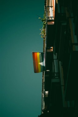 Aşağıdan mavi gökyüzü manzaralı apartman ve balkon parçalarıyla dikey fotoğraf. Balkonda gökkuşağı renkli bayrak Gurur ayında.