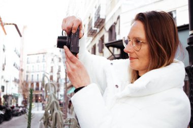 Madrid, İspanya. 1 Mart 2024. Beyaz kürklü genç bir kadın şehir caddesinde küçük bir dijital kamerayla fotoğraf çekiyor. Kadın seyahat fotoğrafçısı..