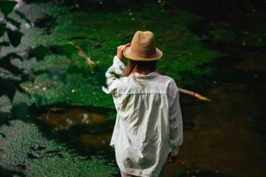 Turist kız yaz ormanlarında yeşil bir göl kıyısında doğal malzeme giysileri ve hasır şapka giyer. Doğadaki sarışın kadın. Doğa dünyasını keşfetmek.