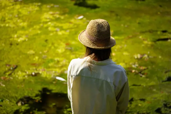 stock image Tourist girl wear natural material clothes and straw hat on a green lake shore in summer forest. Blonde woman on nature. Discover the nature world.