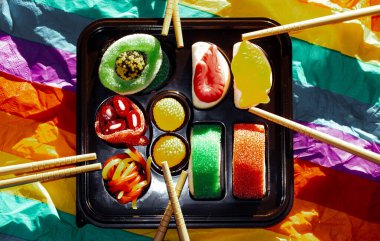 Renkli gökkuşağı arka planında siyah bir kutuya konmuş jöleli şekerler. Tahta suşi çubukları. Çin yemeği. Tatlı şekerler. Neşeli parti yemekleri.