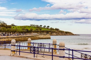 Küçük dalgalı güzel bir deniz manzarası. Baharda bir gezinti güvertesinde, mavi metal çitlerin arkasında deniz kıyısı. Ufukta yeşil tepeler. Demirden korkuluklar.