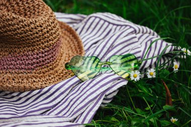 Yeşil kalp şeklinde güneş gözlüğü ve hasır şapka çizgili bir kumaş üzerinde beyaz papatyaları olan yeşil bir çayırda. Yaz mevsimi hala hayattadır. Yaz tatilleri