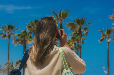 Genç bir kadın yaz mevsiminde bir deniz kıyısı ve palmiye ağaçlarının fotoğrafını çekiyor. Yaz tatilinde bir kız, kameralı bir gezgin. Tropik bölgelere bir gezi.. 