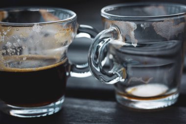 İki küçük, berrak bardak, bir bardak boş ve diğer yarısı da tahtadan bir masanın üzerinde siyah espresso kahvesi. Sıcak sabah içkisi, içecek. Mavi tonlar.