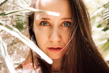 Palmiye yapraklarının arasında esmer bir kadının portresi. Yeşil yaprakların arkasında bir kız. İnsan ve doğa. Düz bakışlı kadın turist tropikal bahçede