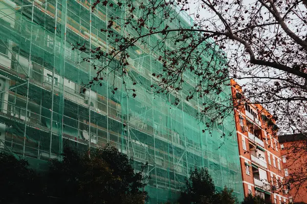 stock image Reconstruction of facade of a historic apartment building on a city street. Green facade construction mesh covers an old house closed for renovation.