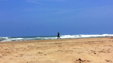 Uzun siyah elbiseli yalnız bir kadın deniz kıyısında yürüyor. Yazın okyanus kumsalında yalnız bir figürle sanatsal bir video. Leylak ve mavi gökyüzü.