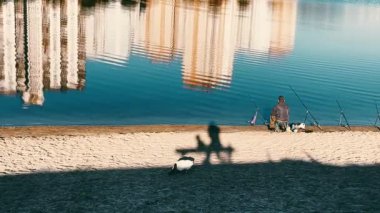 Kyiv, Ukrayna. 25 Eylül 2024 Bir balıkçı ve bir kedi göl, göl, nehir kıyısında. Apartman binaları sakin su yüzeyine yansıyor. Şehir doğası