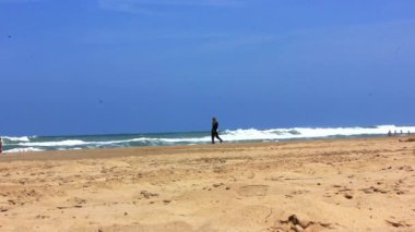 Uzun siyah elbiseli yalnız bir kadın deniz kıyısında yürüyor. Yazın okyanus kumsalında yalnız bir figürle sanatsal bir video. Leylak ve mavi gökyüzü.