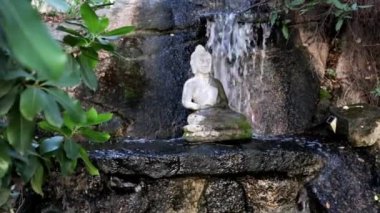 Buda 'nın şelaleye karşı beyaz taş heykeli. Ahenk ve Budizm 'in sembolü. Yeşil yaprakların arasında, Asya bahçesinde meditasyon için bir yer.. 