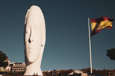 Madrid, İspanya. 22 Kasım 2023 Mavi gökyüzüne karşı gözleri kapalı genç bir kadının dev beyaz heykeli. Colon Meydanı 'nda sanatçı Jaume Plensa' nın başyapıtı. Rüzgardaki İspanyol bayrağı. Hayalperest kız yüzü