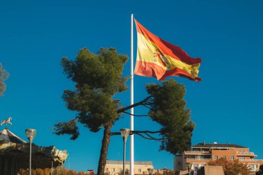 Madrid, İspanya. 22 Kasım 2023 'te bir bayrak direğindeki büyük bir İspanyol bayrağı rüzgârda mavi gökyüzü ve çam ağaçlarından oluşan bir arka planda gelişiyor. Şehir manzarası, kentsel peyzaj evleri çatılar seyahat hedefleri