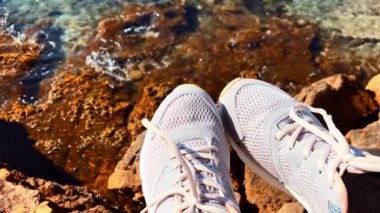 Güneşli bir günde kadın turistin ayaklarındaki gri spor ayakkabılar deniz kenarındaki kayaların üzerinde dinleniyor. Yaz tatili. Yaz tatili dinlenme zamanı..