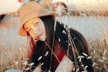 Uzun siyah saçlı, hasır şapkalı güzel esmer bir kadın yaz doğasında rahatlarken eğleniyor. Açık havada bir kadın portresi. Yaz tatili. 