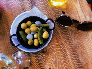 Madrid, İspanya. 1 Ağustos 2023 İspanyol atıştırmalık kasesi yeşil zeytin ve mini salatalıklı, şarap bardağı, bira ve güneş gözlüğü ahşap masa manzaralı..
