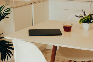 Beyaz bir masada siyah bir laptop ve bir fincan taze sabah kahvesi. Evde çok şık bir çalışma alanı. Kapalı alanda tropik yeşil bölge. Uzaktan çalışma çevrimiçi. 