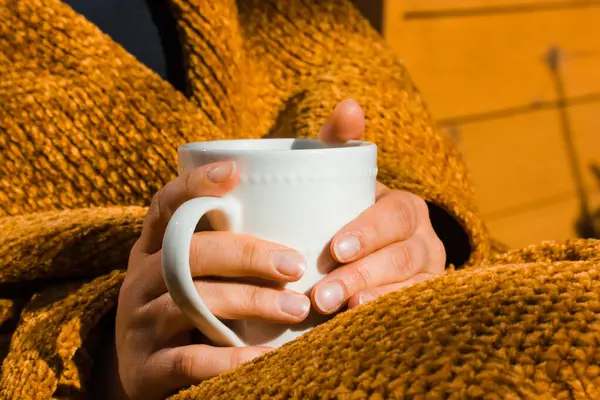 Sonbaharda balkonda oturan bir kadın, elinde beyaz bardakla sarı ekoseye sarılı. Dinlenirken sabah çayı ya da kahve içmek.