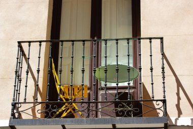 Klasik dövmeli demir balkon, küçük bir masa ve sandalye, güneşli bir dış cephede, eski bir binanın dış kısmında, sarı sandalye ve yeşil masa ile birlikte.