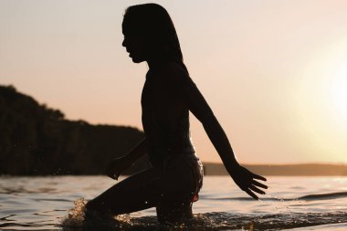 Gün batımında suda yürüyen bir insanın silueti huzur verici, anımsatıcı bir sahne yaratır. Su, gökyüzünün ılık tonlarını yansıtıyor. Huzurlu bir ortam.