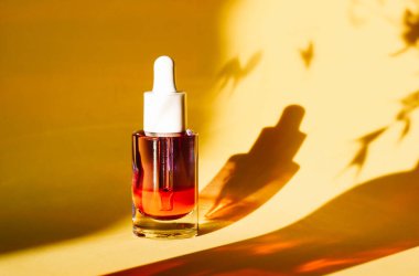 Bir deri serumu şişesi sarı bir yüzeyde duruyor ve yaprakların gölgeleri ılık ve doğal bir ortam oluşturuyor. İçindeki sıvının turuncu-kırmızı rengi var..