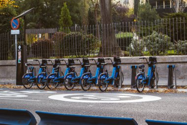 Madrid İspanya 1 Eylül 2024 Bir sıra kiralık bisiklet şehir caddesi boyunca park edilmiş sonbahar ağaçları kullanılmaya hazır. Mavi kiralık motorlar düzgün bir şekilde dizilmiş.